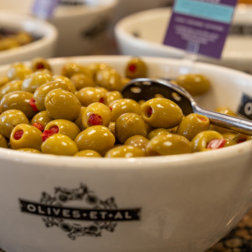 Bowl of olives with a spoon, branded 'Olives et al', on a table.