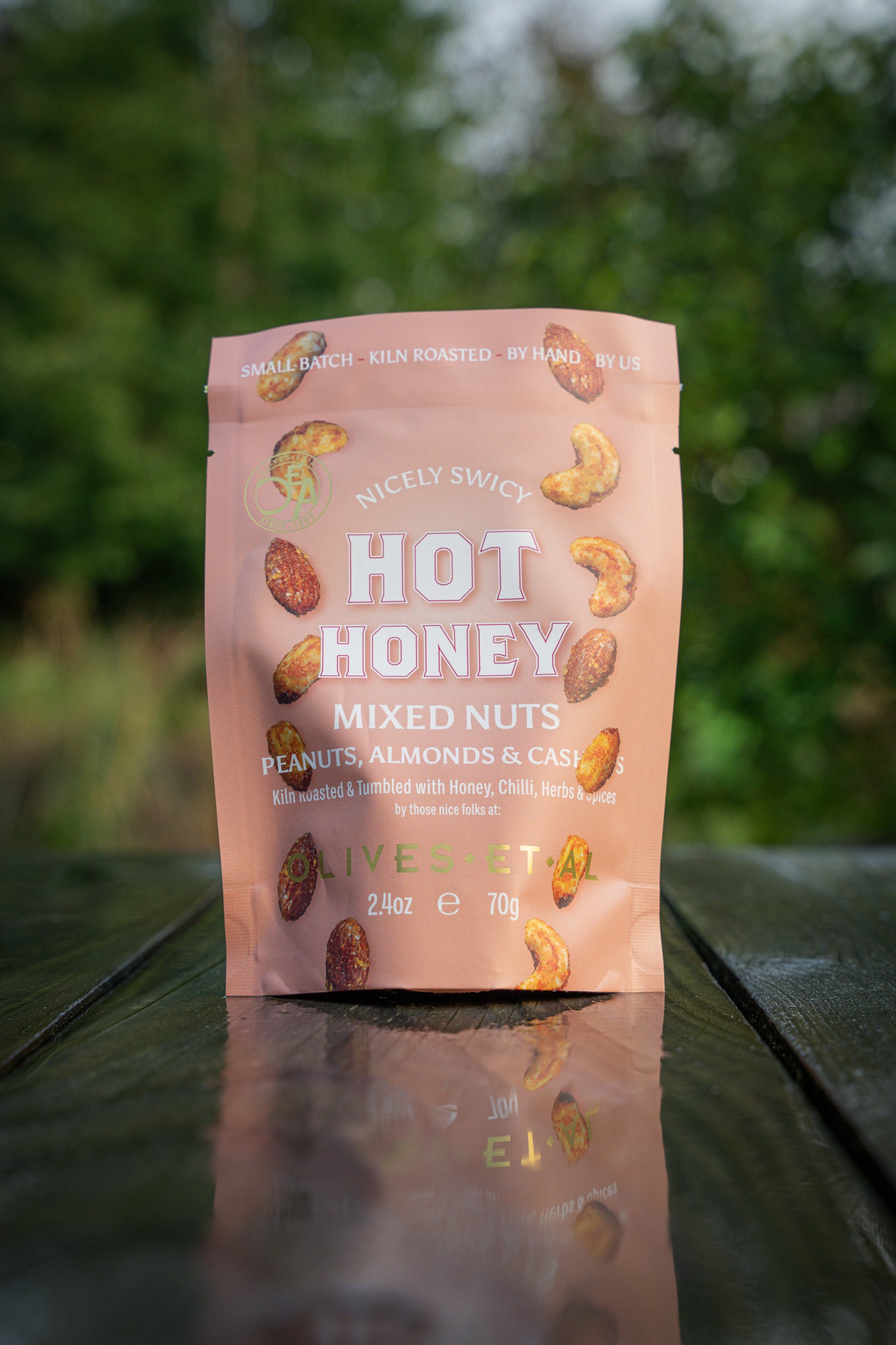 Pink package of Nicky Snacky Hot Honey Mixed Nuts on a reflective surface with a blurred green background