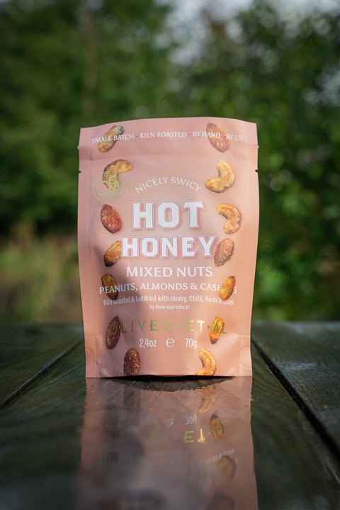 Pink package of Nicky Snacky Hot Honey Mixed Nuts on a reflective surface with a blurred green background