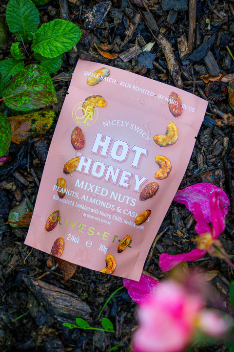 Pink packet of Nicely Sweet Hot Honey Mixed Nuts on a natural background with plants and flowers.