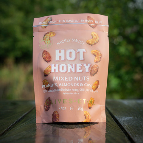 Packaged mixed nuts with 'Hot Honey' flavor on a reflective surface with a blurred natural background