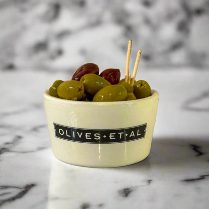 A ceramic bowl containing pitted green and Kalamata olives with fennel and orange, branded with 'OLIVES.ET.
