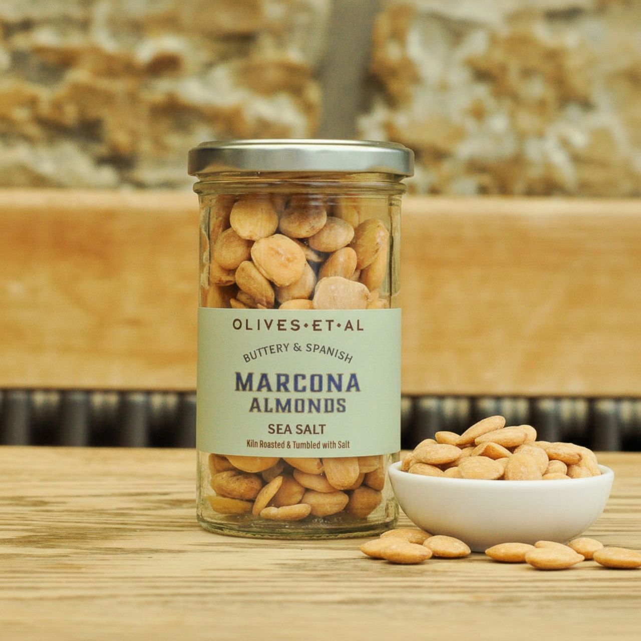 Olives Et Al Jar of Olives et al Marcona Almonds with a bowl of almonds on a wooden surface.