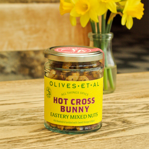 Jar of 'Hot Cross Bunny' mixed nuts with a yellow label on a wooden surface, accompanied by yellow flowers.