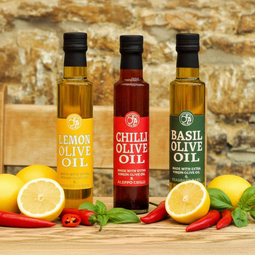 Three bottles of olive oil with lemon, chili, and basil on a wooden surface against a stone wall background.